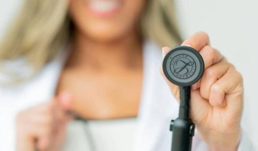 Smiling healthcare professional holding black stethoscope, white coat visible, medical supplies
