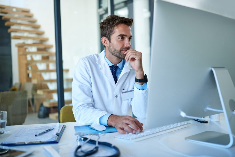 Doctor in white lab coat working at computer in modern medical office with stethoscope on desk