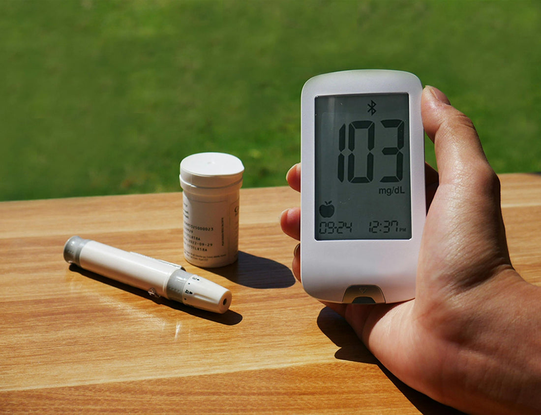 Hand holding blood glucose meter showing 103 mg/dL with test strips and lancet on wooden table