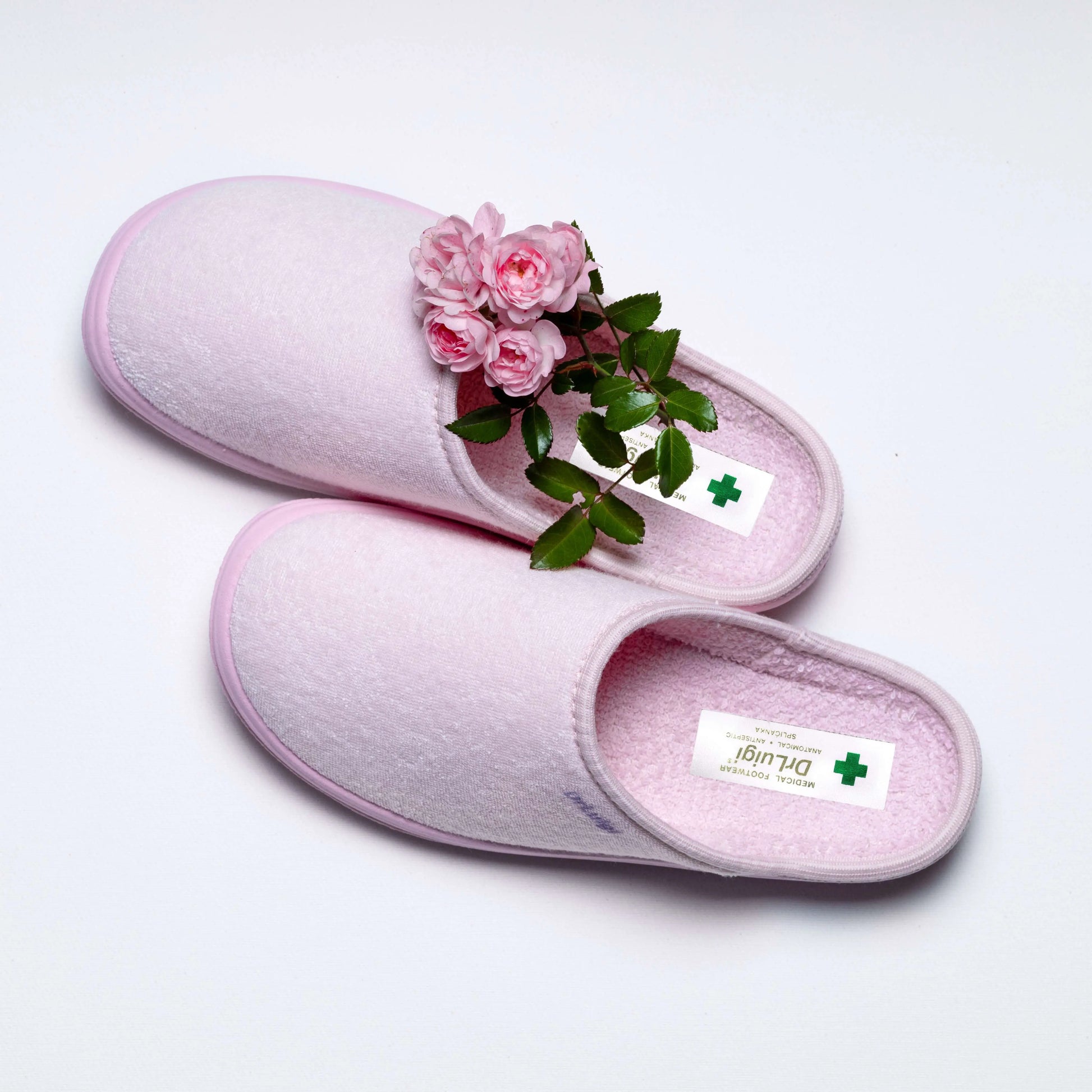 Pair of pink medical slippers with green cross tag and fresh pink roses on white background